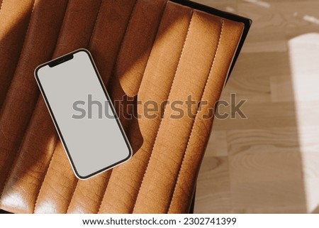 Flatlay mobile phone on leather bench or chair with elegant soft sunlight shadows. Flat lay, top view. Aesthetic copy space mockup template