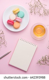 Flatlay Of Notebook, Cake Macaron, Cup Of Tea And Flower On Pink Table. Beautiful Breakfast With Macaroon.