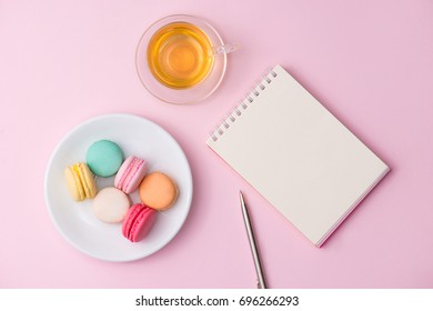 Flatlay Of Notebook, Cake Macaron And Cup Of Tea On Pink Table. Beautiful Breakfast With Macaroon.