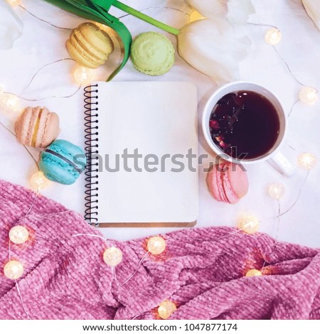 Flatlay of notebook, cake macaron, cup of tea and glowing garland on a background of a pink knitted blanket. Beautiful breakfast with macaroon.
Plans for the day.