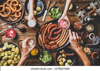 Flat-lay of Octoberfest party dinner table with grilled meat sausages, German pretzel pastry, potatoes, cucumber salad, sauces, beers and peoples hands with food over dark wooden background, top view