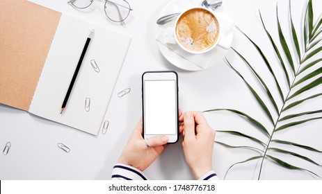 Flatlay on white work space desk with woman's hands holding white smartphone with white copyspace, coffee, planner and other office accessories. Mobile phone mockup.