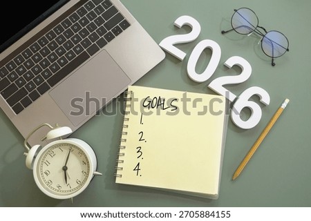 flat-lay of open notebook labeled “Goals” with lists of thing to do, number of New Year 2026, pencil, laptop, alarm clock and eyeglasses on green desk. goal setting, planning for New Year concept