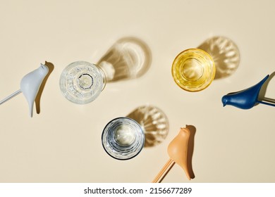 Flatlay Of Three Glasses Containing Water, Their Shadows And Small Toy Birds Of Various Colors Over Beige Background Or On Table