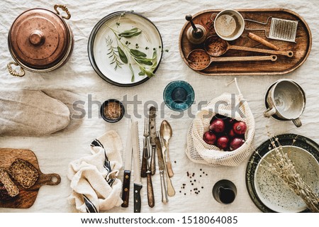 Flat-lay of various kitchen utensils, rustic tablewear, plates, dishes, glasswear, pan, mitten, fresh bread and seasonak red onion over white rustic linen tablecloth background, top view