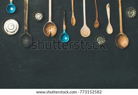 Flat-lay of various old vintage kitchen spoons and baking tin molds over black stone background, top view, copy space. Rustic cooking concept
