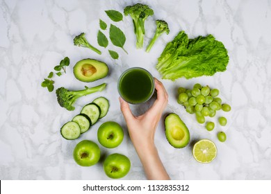 Flatlay With Woman's Hand Reaching For Green Juice Surrounded By Green Fruits And Vegetables On Marble Table