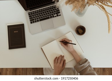 Flatlay of women's hands writing in a notebook sheet. Comfortable home office workspace interior design. Work at home. Laptop computer, notebook, pampas grass bouquet on table. Flat lay, top view. - Shutterstock ID 1917250406
