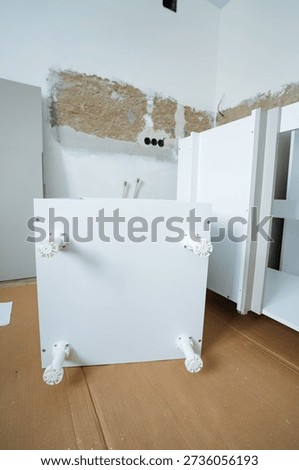 FlatPacked Cabinet Ready For Installation On Cardboard Floor Protection, UpsideDown Orientation Showing Legs And Mounting Points Staged Assembly Scene During Kitchen Renovation Preparation.