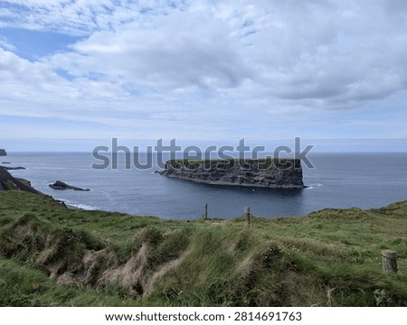 A flat-topped sea stack off the rugged coast of Ireland, viewed from a grassy clifftop pasture with a traditional wooden fence.
