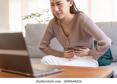 Flatulence asian young woman hand in stomach ache suffer from food poisoning, gastritis or diarrhoea, pain belly, abdomen or inflammation, use laptop computer consulting with doctor online at home. - Shutterstock ID 2049644216