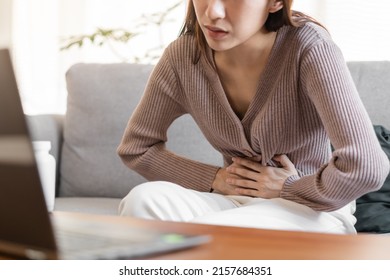 Flatulence asian young woman hand in stomach ache suffer from food poisoning, gastritis or diarrhoea, pain belly, abdomen or inflammation, use laptop computer consulting with doctor online at home. - Shutterstock ID 2157684351