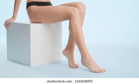 Flawless female legs. Young slim woman in black underwear sitting on the white cube platform on pale blue background | Leg care and unwanted hair removal concept