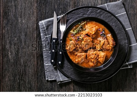 Flemish Stew, stoofvlees, carbonnade, beef or pork, beer and onion stew in black bowl with cutlery on dark wood table, horizontal view from above, flat lay, free space