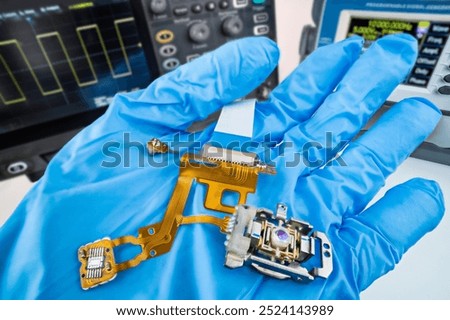Flex printed circuit, sensor or lens of CD-DVD disc drive pickup head. Electronic components and ribbon cable on expert hand in technical lab at adjusting with oscilloscope and DDS function generator.