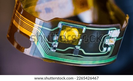 Flex printed circuit and small electronic components in plastic strip curled into circle on dark background. Closeup of ribbon cable with green and yellow copper lines on flexible PCB from headphones.