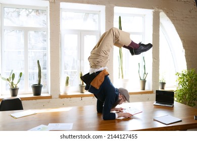 Flexibility. Man practicing yoga. Young bearded office clerk having fun, doing yoga on wooden table in modern office at work time with gadgets. Concept of business, healthy lifestyle, sport, hobby - Shutterstock ID 2144127455