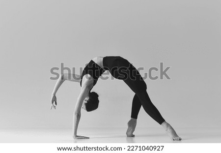 Flexibility and strength. Black and white photography. Young girl doing stretching exercises over studio background. Concept of sport, body care, beauty, fitness, active lifestyle. Ad