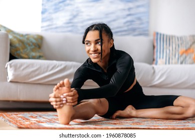 Flexible active attractive African American girl dressed in sportswear, engaged in fitness, leads a healthy lifestyle, stretches on the floor at home, stretching out her arms to the foot, smiling - Powered by Shutterstock