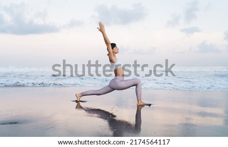 Flexible Caucasian trainer in stylish tracksuit spending weekend morning for stretching at coastline beach enjoying healthy lifestyle, charming female exercising at seashore doing pilates warm up