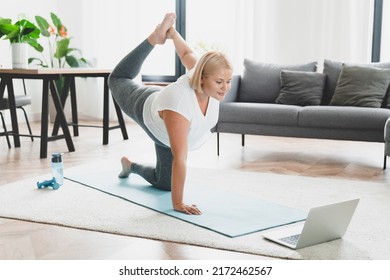 Flexible caucasian young athlete plus size plump woman training at home, stretching her leg watching online video tutorial on laptop. Shaping slimmimg on fitness mat