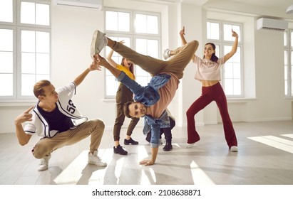 Flexible And Energetic Male Dancer Has Fun With His Dance Group Doing Elements Of Breakdance Movements. Young People In Stylish Youth Casual Clothes Dance In Bright Dance Hall With Windows.