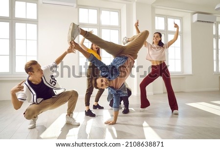 Flexible and energetic male dancer has fun with his dance group doing elements of breakdance movements. Young people in stylish youth casual clothes dance in bright dance hall with windows.
