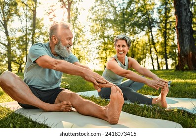 Flexible exercises for body. Sporty man and woman with grey hair stretching on yoga mats with hands to one leg during outdoors workout. Happy married couple with bare feet warming up together at park. - Powered by Shutterstock