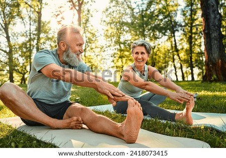 Flexible exercises for body. Sporty man and woman with grey hair stretching on yoga mats with hands to one leg during outdoors workout. Happy married couple with bare feet warming up together at park.