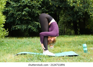 Flexible, slim young woman in tight sportswear practicing yoga asana uttanasana on sports mat on green grass in park. Side view caucasian athletic sportswoman stretching her spine, exercising outdoors
