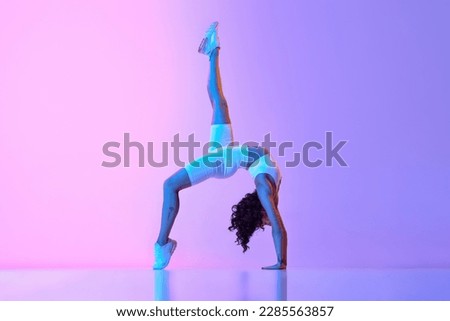 Flexible, sportive young woman training, doing stretching exercises against gradient pink blue background in neon light. Concept of sportive lifestyle, beauty, body care, fitness, health