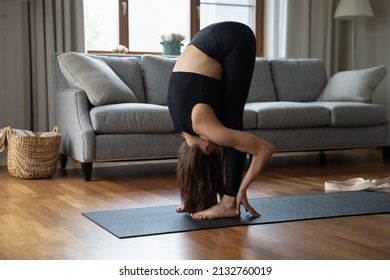 Flexible woman in black sportswear practicing yoga, standing on mat do forward bend exercise or Head to Knees, Uttanasana pose, working out alone at modern home, stretches hips and hamstrings concept