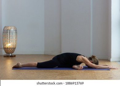 Flexible Woman Lying Down On Pigeon Pose At Yoga Studio. Female Yogi On Black Outfit Performs Sleeping Swan With One Leg Stretched, The Other One Folded And Arms Forward. Wellbeing Concept