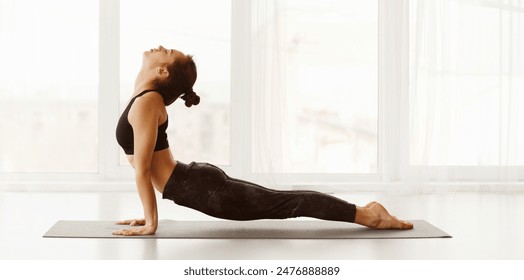 Flexible woman performs an upward-facing dog pose in a well-lit yoga studio. She is focused on her posture as natural light fills the room, creating a serene and peaceful atmosphere, panorama - Powered by Shutterstock