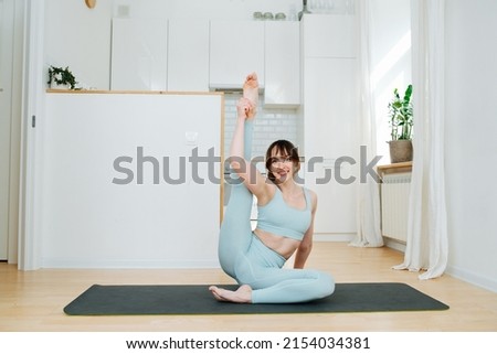 Flexible woman rasing her leg next to her head on a mat. In a large spacious kitchen. Wearing blue sportswear.