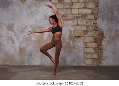 Flexible young dark skinned ballet dancer with curly brown hair stretching over brick wall background, wearing sporty black top and brown leggins, having earphones in her ears - Powered by Shutterstock