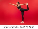 Flexible young woman in black sportswear exercising on red studio background, practicing yoga or pilates, full length