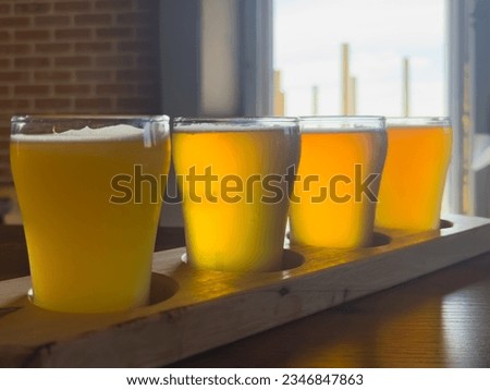A flight or an assortment of craft beers in small clear glasses on a wooden paddle in a restaurant pub. Each of the tasting sample beers ranges from dark yellow to golden color with froth on top. 