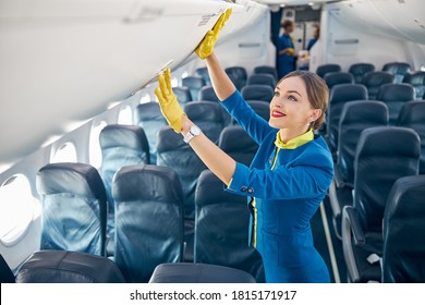 Flight Attendant In Blue Suit And Yellow Lather Gloves Checking Compartment With Hand Luggage While Standing In The Empty Board Of Commercial Airplane