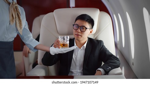 Flight Attendant Giving Glass Of Whiskey To Asian Businessman In Suit In Private Plane. Stewardess Serving Scotch To Japanese Male Executive Flying First Class