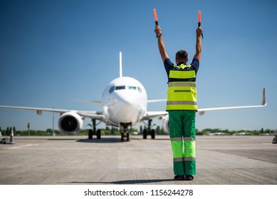Flight is over. Back view of airport worker meeting passengers and directing the plane. Blue sky, aircraft and runway on background