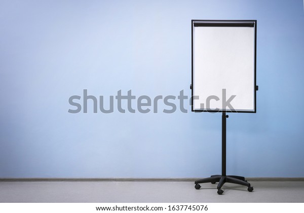 Flip chart board on wheels with a blank sheet\
of paper on a light blue wall background, copy space. Empty room\
for educational\
activities.