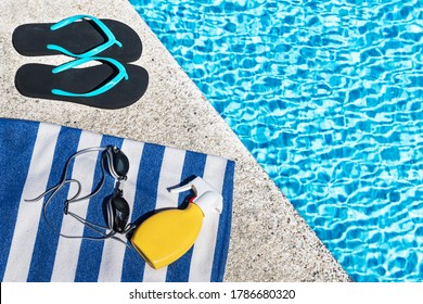 Flip Flops, Sunscreen Spray And Swimming Goggles On A Blue And White Striped Towel At The Edge Of A Pool.. Copy Space. Top View.