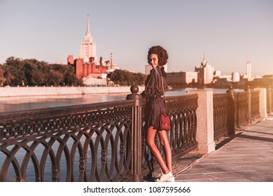 Flirtatious charming young black tourist girl with a curly afro hair is leaning on the fencing of the embankment next to Moscow river and smiling, with the hight-rise in a defocused background - Powered by Shutterstock