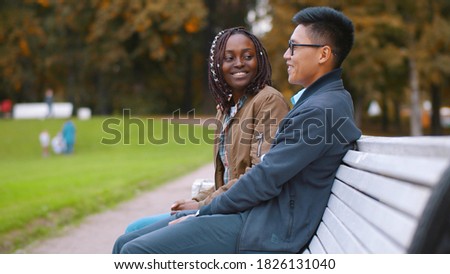 Flirting diverse couple sitting on bench in park spending time together. Young happy chinese man and african woman relaxing on bench outdoors and talking enjoying romantic date