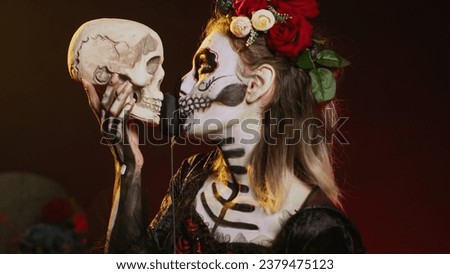 Flirty dead lady acting creepy with skull in hand, holding black roses and wearing scary make up. Looking like santa muerte with body art on holy mexican celebration, being charming. Handheld shot.