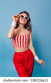 Flirty Woman Of 80s Is Smiling Sweetly Against Blue Background. Lady In Striped Top And Red Pants Puts On Sunglasses