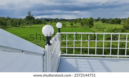 The floating balcony on the second floor, close to the roof of the house, is equipped with a white round lamp with a black base pole. To use as a view point of the next green rice field. 