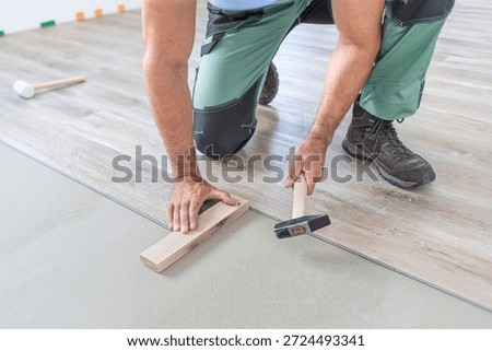 Floating floor work. The worker taps the board of vinyl plank to lock the click system. He is using hammer and wooden plank.