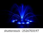 Floating fountain in neon blue, large detailed colorful horizontal closeup at night, lit up illuminated color changing bright city park lights, isolated colored water jets on dark black background